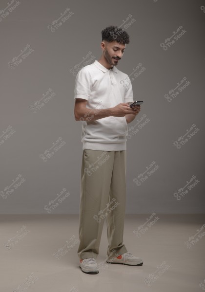A young man standing in a studio with a grey background, wearing a white polo shirt and beige trousers with white sneakers. He appears to be using a mobile phone and is looking at it with a slight smile.