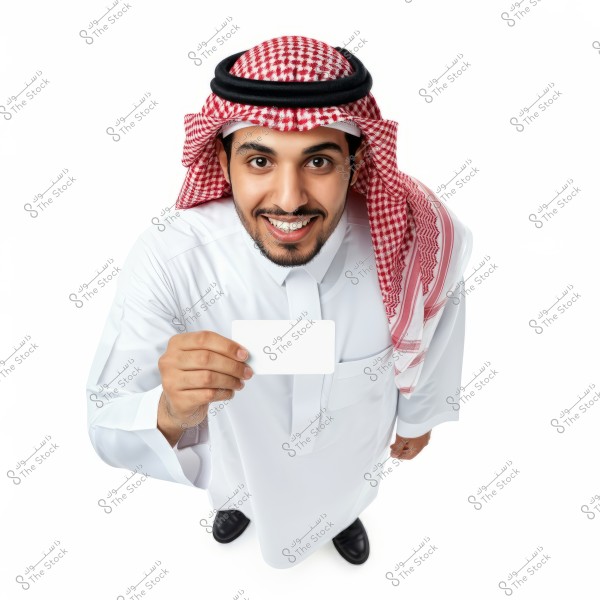 An image of a man wearing a white thobe and a red and white checkered ghutra with an agal. He is smiling and holding a blank white card. The photo is taken from a high angle. The attire is traditional Gulf style, likely from Saudi Arabia.