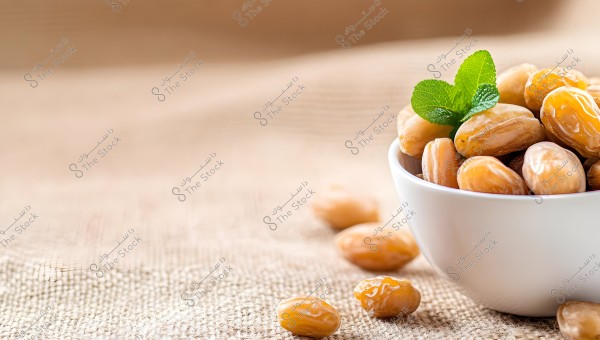 An image of a white bowl containing brown dates with a green mint leaf on top, placed on a piece of burlap. A few dates are gently scattered around the bowl.