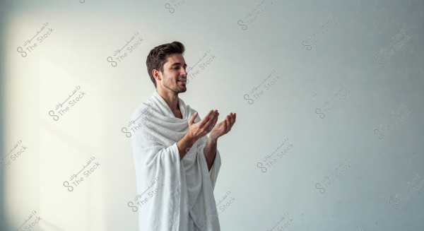 An image of a man wearing a white ihram, seemingly engaged in prayer or supplication. The background is empty with an off-white to gray color, highlighting the person in the image.