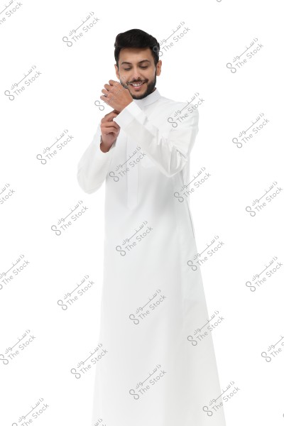 Image of a man wearing a white thobe, smiling and adjusting his cufflinks. He stands against a white background, highlighting the classic look of traditional attire. The thobe suggests the image might be inspired by the Gulf region.