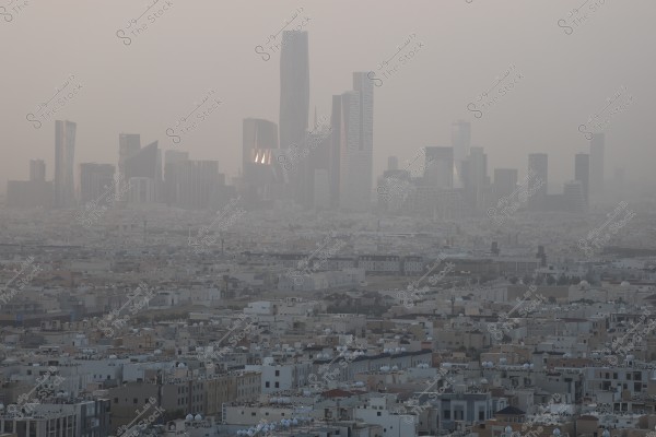An aerial view of a modern city with several skyscrapers rising above a foreground of low-rise residential buildings. The skyline appears shrouded in a layer of dust or haze, adding a mysterious appearance to the scene. The towering buildings stand out with their contemporary architectural designs, likely situated in a city with a desert climate.