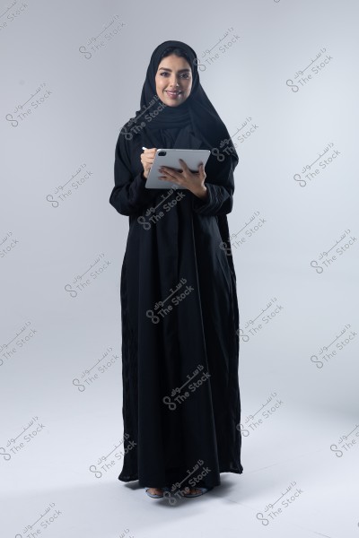 An image of a woman wearing a black abaya and hijab, standing with a smile while holding a silver tablet. The woman appears to have Middle Eastern features, possibly from Saudi Arabia. The background is white, highlighting the details of her clothing.