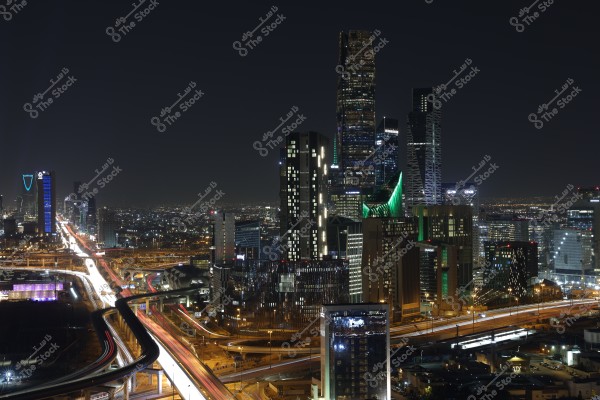 A nighttime photo of Riyadh city in Saudi Arabia showcasing an array of illuminated skyscrapers. The Al Faisaliah Tower is visible in the background on the left, and the modern architecture of the Riyadh skyline is prominent in the center, lit by bright lights. Streaks of cars are visible on the highways weaving through the buildings.