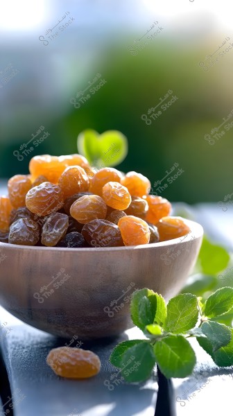 An image of a wooden bowl filled with brown and golden raisins on an outdoor surface, surrounded by green leaves. The background is blurred and bathed in bright sunlight.