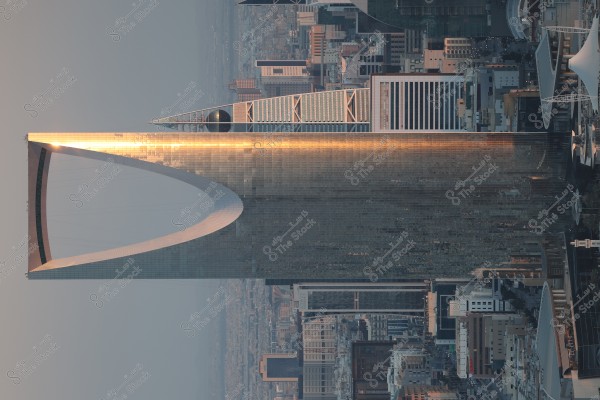صورة لبرج المملكة في الرياض, عاصمة المملكة العربية السعودية. يظهر البرج الشاهق بتصميمه المعماري الفريد الذي يتألف من قوس في الأعلى يتوسطه جسر. تحيط به العديد من المباني العالية الأخرى في وسط المدينة, وتنعكس أشعة الشمس على الهيكل الزجاجي.