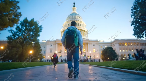 A man wearing a gray hoodie and jeans with a green backpack is walking towards the Capitol building in Washington, D.C. The building is lit in the evening, surrounded by trees and greenery. Street lights are on, and the sky is a clear blue at sunset.