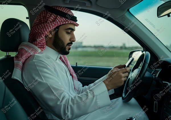 A man wearing a white thobe and a red ghutra is seated inside a car, using a mobile phone. The man appears to be of Arabian heritage, possibly from Saudi Arabia, as he is wearing traditional Saudi attire.