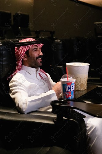 شاب سعودي في صالة عرض  السينما , يرتدي الزي الرسمي السعودي ، يظهر امامه فشار و مشروب