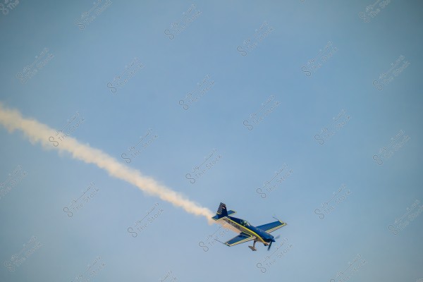 An aerobatic blue and yellow plane flying in the clear sky with a white smoke trail behind it. The plane is performing a maneuver and is tilted in the air.