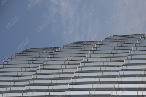 An image depicting part of a building with reflective glass panels mirroring the blue sky. The exterior design consists of successive horizontal layers arranged symmetrically, adding a modern touch to the structure. The sky appears in the background with a slightly cloudy blue backdrop.