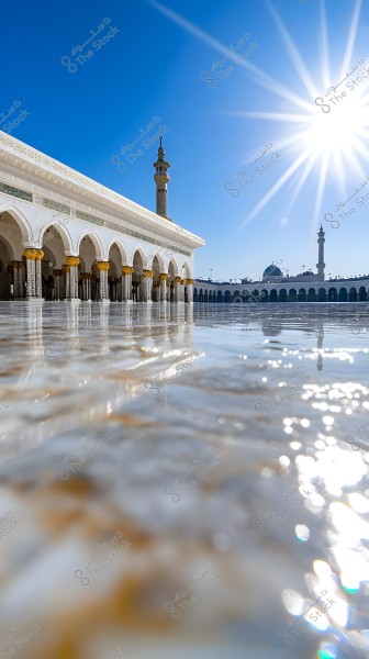 A view of a mosque complex with sunlight reflecting off the shiny marble floor. In the background, a section of the clear blue sky with the bright sun and a corner of Islamic architectural buildings with domes and minarets are visible.