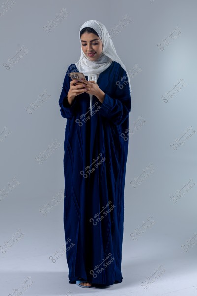 Image of a woman wearing a dark blue abaya and a white hijab, standing while looking at a mobile phone in her hands, set against a simple gray background.
