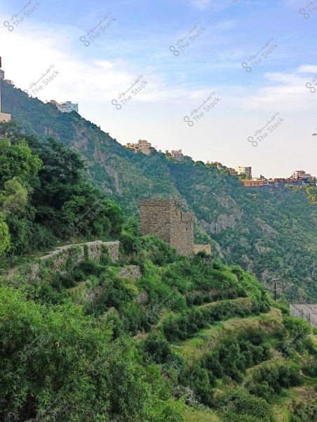A natural landscape showing green mountainous terrain with agricultural terraces. Some buildings are visible on top of the mountain, while remnants of an old stone structure are visible in the foreground. The sky is blue with light clouds.