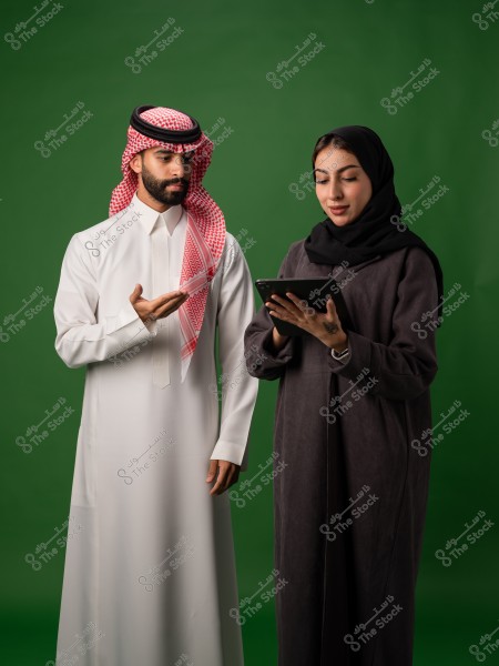 رجل يرتدي الثوب السعودي التقليدي يقف بجانب امرأة محجبة ترتدي عباءة سوداء تحمل جهازًا لوحيًا, وخلفهم خلفية خضراء.