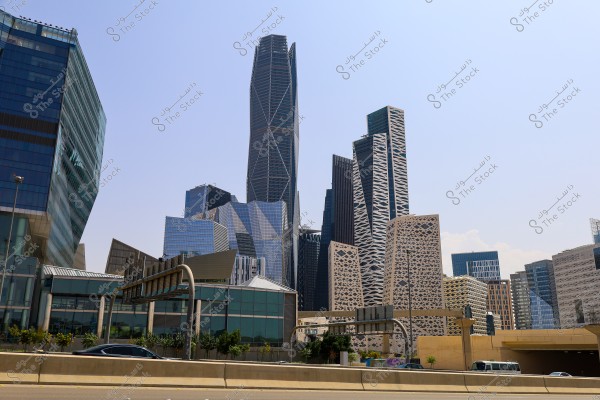 View of a group of modern skyscrapers in Riyadh, Saudi Arabia. The buildings feature innovative and diverse architectural designs with glass facades and geometric patterns. A highway is seen in the foreground with cars passing by. The sky is clear and almost cloudless.