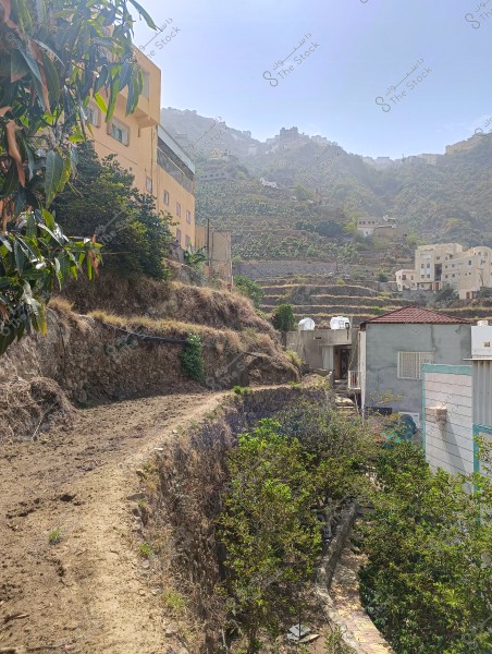 Image depicting a mountainous scene with terraced agricultural fields and several houses. The picture shows light-colored buildings scattered across the slopes, with green terraces descending downwards. The sun is bright and the sky is generally clear in the background.