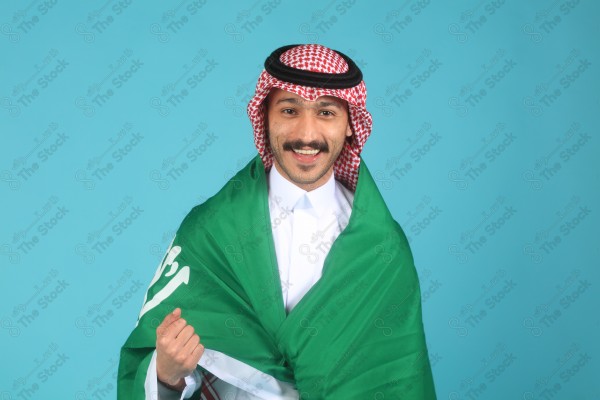 A young Saudi man wearing the Saudi dress and holding the Saudi flag makes expressions of happiness and enthusiasm،Saudi flag