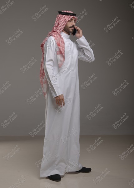 A man wearing traditional Saudi attire is speaking on the phone. He is dressed in a white thobe with a red and white checkered headscarf (ghutra) and a black headband (agal). He is standing against a gray background and is shown from the side.