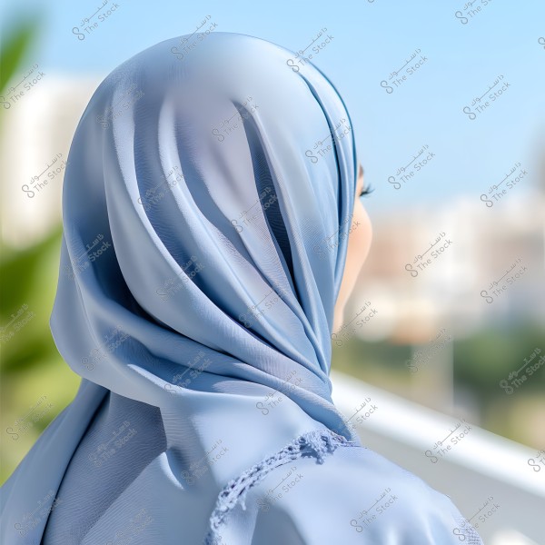 An image showing a woman wearing a light blue hijab, viewed from behind as she stands in a sunny outdoor setting. The background is blurred and includes some greenery and buildings.