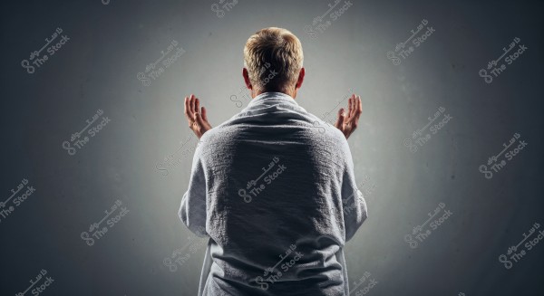 Image of a man shown from behind wearing a white garment, appearing in a posture of prayer with his hands raised. The background is a simple gray, highlighting the details of the white garment.