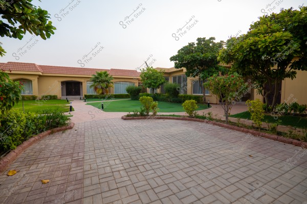 An image of a large house courtyard featuring a tiled pathway splitting in two directions, surrounded by green lawns, trees, and plants. The house has a sloped roof covered with red tiles and large windows. The clear sky suggests a tranquil and sunny atmosphere.
