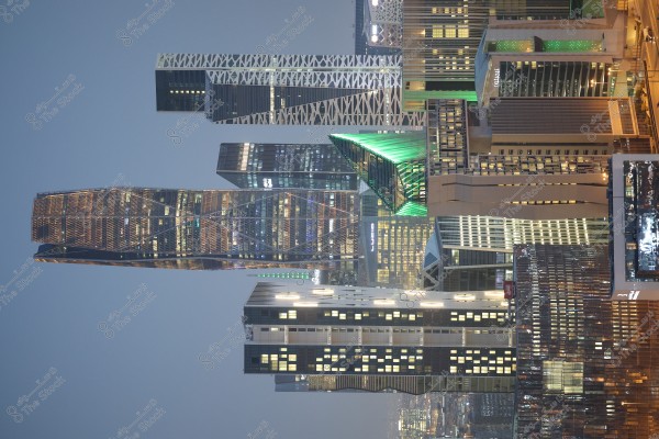 Image showing a cityscape at dusk with a collection of modern towers in the skyline. Green lights illuminate one of the buildings, highlighting its distinct architectural design. The sky is a light blue, blending with the reflected lights from the buildings, creating a bright and vibrant scene.