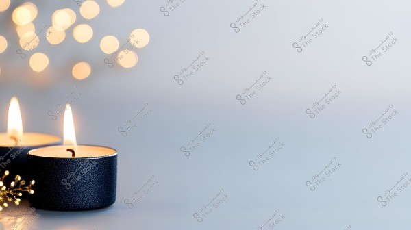 Two small black candles are lit with a gentle glow on a gray surface. The background features warm bokeh lights, adding a cozy and calming ambiance to the image.