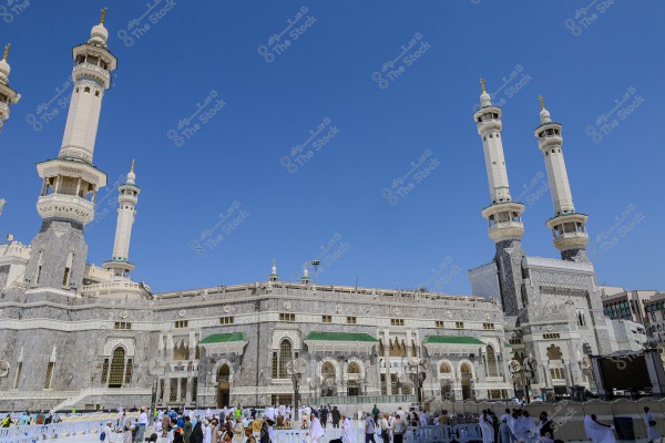 The image shows the Holy Mosque (Masjid al-Haram) in Mecca, Saudi Arabia. Several white minarets with ornate domes stand tall against a clear blue sky. Many worshippers wearing white garments, indicative of the Ihram, are gathered in the mosque\'s courtyard.