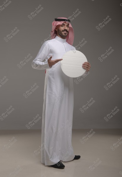 A portrait of a man standing in a studio wearing traditional white thobe, ghutra, and agal, suggesting Gulf region origin. He is holding a circular board and pointing at it with his other hand. The background is a neutral gray tone.