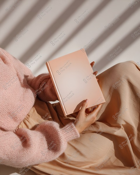 A woman wearing a pink knitted sweater and beige skirt holds a closed light pink book. Light and shadow from a window create a pattern on the white background.