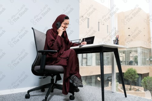 A Saudi woman wearing an abaya works in a glass-fronted office and uses a mobile device to write while taking notes during the day