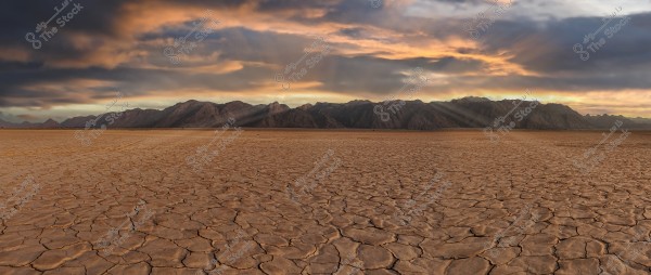 A natural landscape of a barren desert with cracked earth extending to mountainous terrain in the background. The sky is filled with clouds colored in orange and blue, reflecting the setting sun.