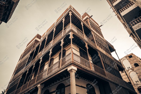 صورة لمبنى تقليدي قديم ذو طراز معماري عربي في منطقة تاريخية. يتميز المبنى بشرفاته الخشبية المزخرفة والنوافذ الكبيرة المغطاة بألواح خشبية. يظهر المبنى بارتفاعه الشاهق وتفاصيله المعمارية المميزة تحت سماء دافئة مضيئة.
