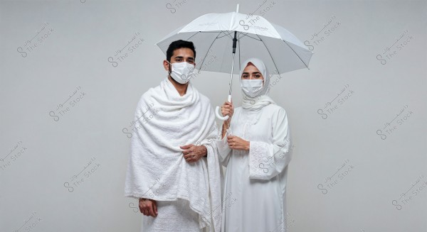 The image shows a man and a woman wearing white Ihram clothing, standing next to each other. The man is wearing a white Izar and Rida, while the woman is dressed in a long white dress with a headscarf. Both are wearing white face masks, and the woman is holding a white umbrella. The background is a plain grey.