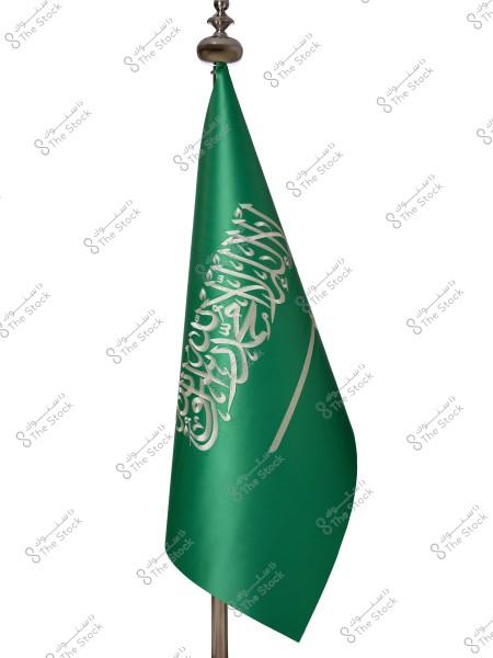 The flag of Saudi Arabia displayed on a flagpole against a white background. The flag is green with white Arabic inscription of the Shahada, and a white sword below it extending horizontally.