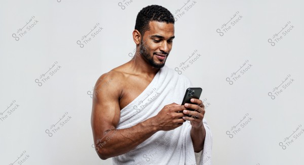 Image of a man standing, wearing a white towel draped over his shoulder and part of his body, while looking at a smartphone in his hand and smiling. The background is plain white, highlighting the focus on the person in the image.