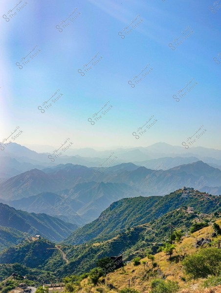 صورة لمنظر طبيعي خلاب سُحِبَت في منطقة جبلية تظهر فيها سلسلة من الجبال الخضراء الممتدة في الأفق. السماء زرقاء وصافية, توفر خلفية هادئة للمشهد. يمكن رؤية بعض المنازل الصغيرة المتناثرة على تلال الجبال في مقدمة الصورة.