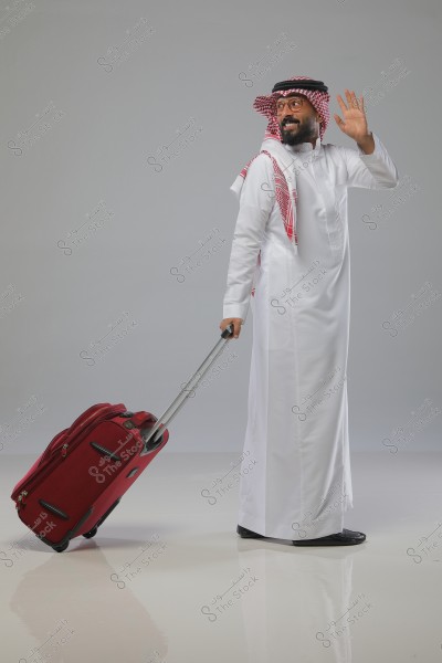 A man dressed in a white thobe and traditional headscarf waves his hand while pulling a red suitcase. He stands on a reflective gray floor.