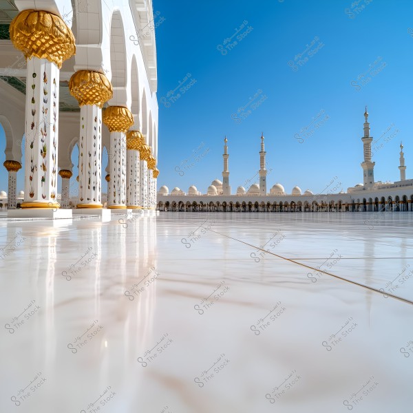 An image showing the courtyard of a large mosque with ornate columns topped with golden capitals. The mosque\'s white, glossy backdrop features numerous domes and tall minarets under a clear blue sky.
