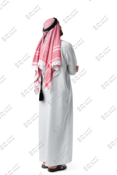 Image of a man wearing a white thobe, viewed from the back. The man is wearing a red and white checkered shemagh styled traditionally and secured with a black agal. He is wearing black sandals and is standing against a neutral white background.