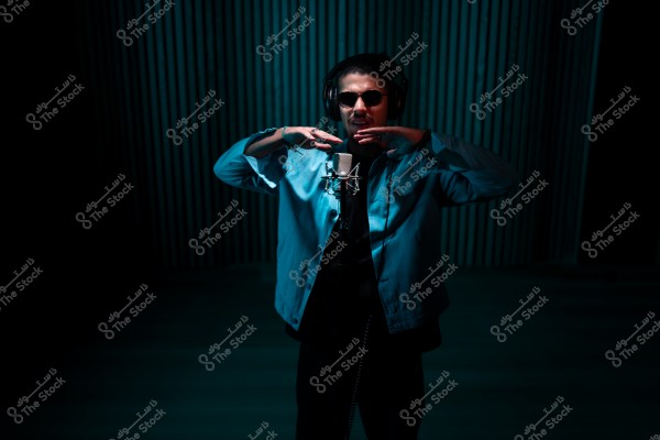 شخص يرتدي سماعات رأس ونظارات شمسية يؤدي في استوديو تسجيل مضاء بإضاءة خافتة.