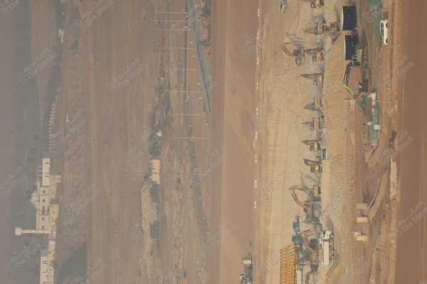 The image shows a large construction site in a desert area. The site features several excavators and heavy machinery working on the ground. There is also a row of light poles extending horizontally, with low buildings and a long fence visible in the background, along with some parked vehicles.