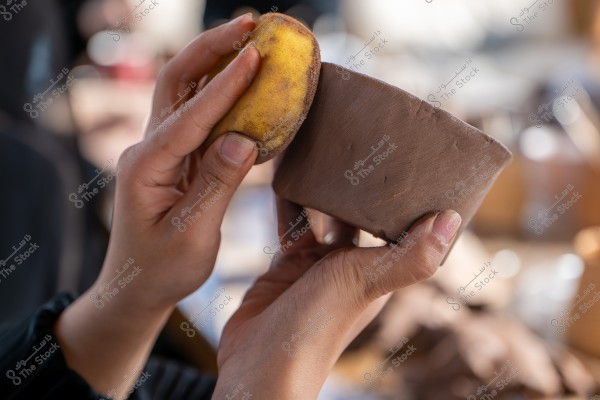 The image shows two hands engaged in pottery making. The left hand holds a damp, semi-circular piece of clay, while the right hand uses a yellow sponge to smooth the surface. The blurred background suggests a crafts workshop.