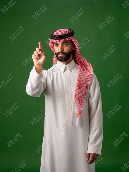 A man wearing traditional Saudi attire stands in front of a green background, raising his finger.