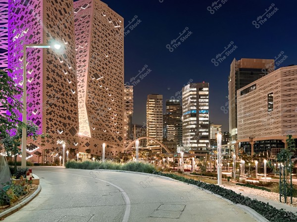 The image shows a wide walkway lit by streetlights at night, surrounded by modern skyscrapers. The buildings feature innovative architectural designs with colorful lighting, with one structure prominently lit in purple. Trees and plants decorate the area surrounding the walkway. The dark sky accentuates the nighttime lighting of the buildings.