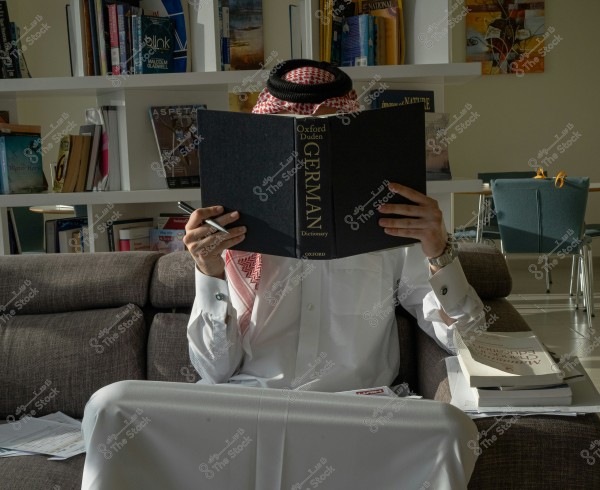 Image of a man sitting on a couch, wearing traditional Saudi attire consisting of a thobe and a ghutra, reading a German language dictionary. Behind him is a bookshelf filled with various books and magazines. Additional books and papers are scattered around him on the couch and nearby table.