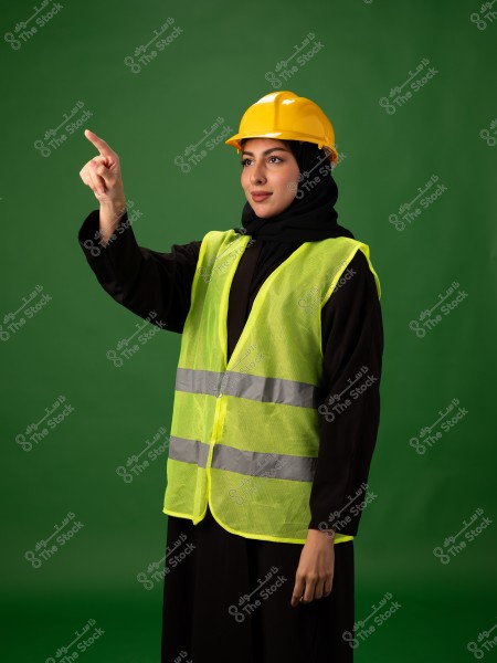 Image of a woman wearing a yellow safety helmet and a yellow reflective vest over a black abaya, set against a green background. She is raising her hand and pointing, suggesting attention or direction. She appears to represent a role related to construction or safety.