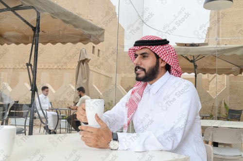 رجل سعودي يرتدي الزي السعودي التقليدي يضع ساعة على معصمه بينما يحتسي كوب من القهوة أثناء النهار