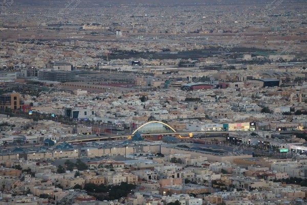 تُظهر الصورة مشهدًا من مدينة الرياض في السعودية, حيث يتم تصوير المدينة من الأعلى موضحاً توزيع المباني وطرق المدينة المزدحمة. يوجد جسر ملون بالقوس في وسط الصورة, مع أضواء تظهر على الطريق وواجهة محلات تجارية.